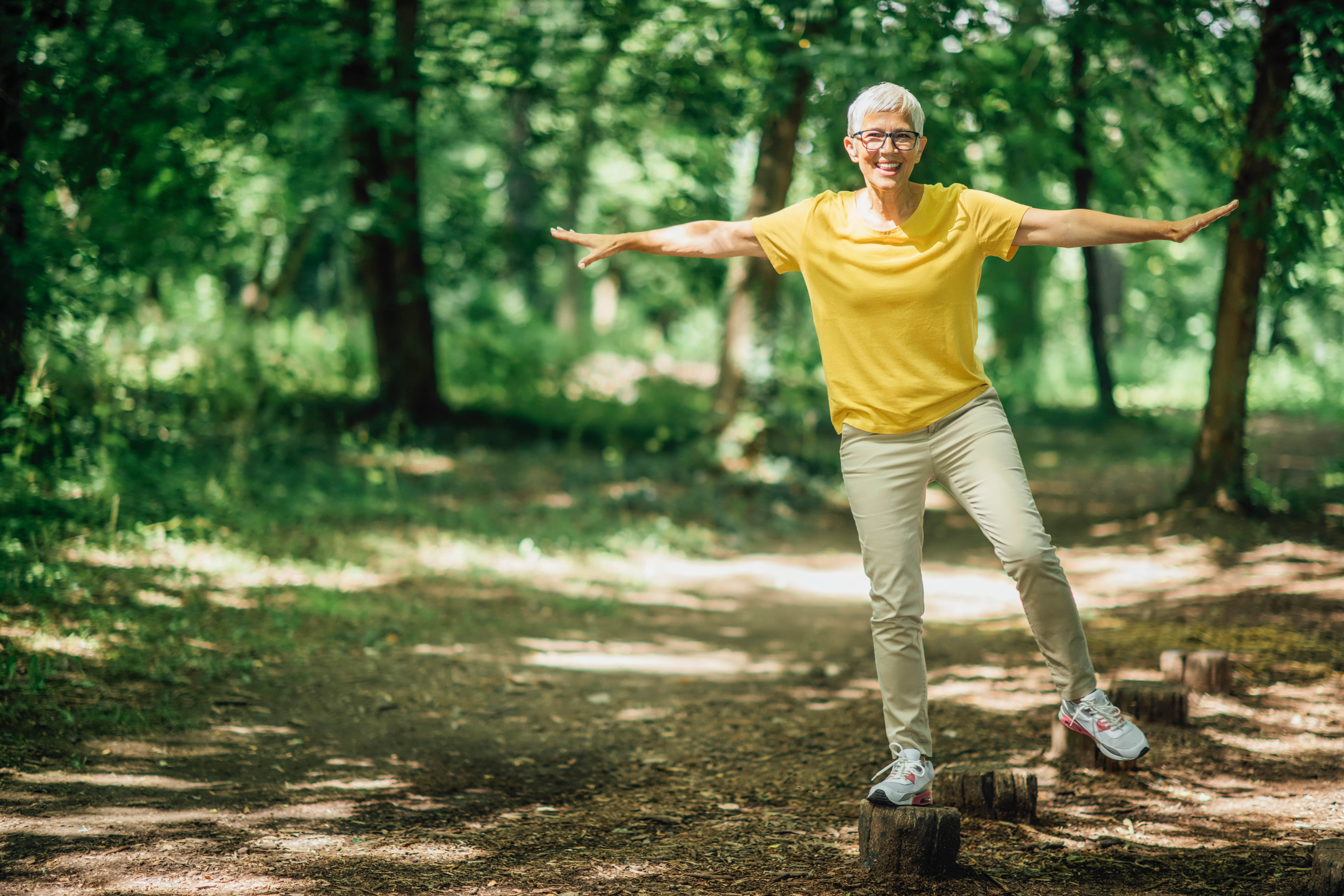 Older woman standing on a tree stump on one foot, doing some balance work outdoors